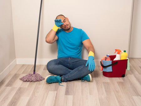 Young man cleaning with a sweeping brush and mop and bucket of water and cleaning supplies at home wearing blue gloves and blue shirtの写真素材