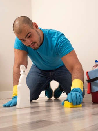 Young man cleaning with a sweeping brush and mop and bucket of water and cleaning supplies at home wearing blue gloves and blue shirtの写真素材
