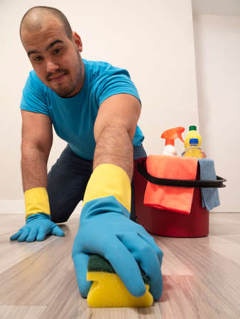 Young man cleaning with a sweeping brush and mop and bucket of water and cleaning supplies at home wearing blue gloves and blue shirtの写真素材