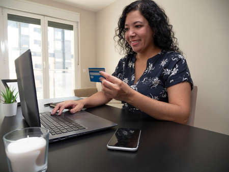Young caucasian woman with deep hair working from her living room on a laptop on a black table with a white candle, her phone and a credit card with a black notebookの写真素材
