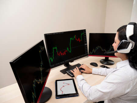 Caucasian young woman sitting at a table doing stock trading on a computer with three black monitors...