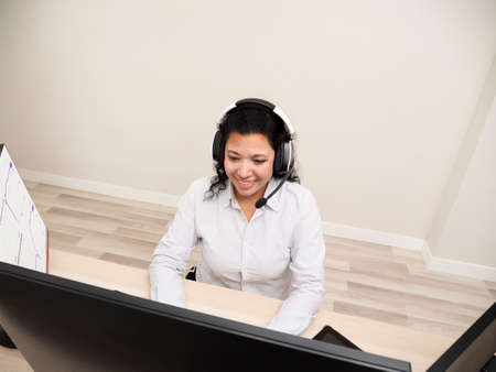 Caucasian young woman sitting at a table doing stock trading on a computer with three black monitors in a white roomの写真素材