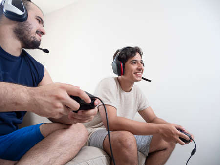 Two young adults playing video games with video game controls on a white sofa in a white room, a...