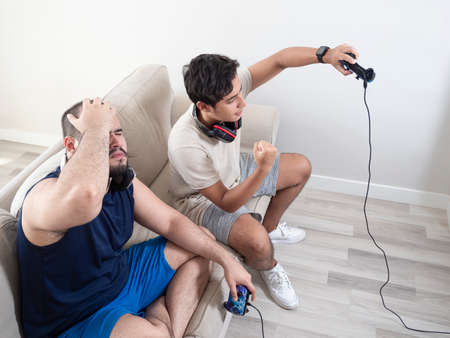 Two young adults playing video games with video game controls on a white sofa in a white room, a Caucasian youth and a dark-skinned oneの写真素材