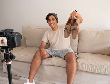 Hispanic young man creating clothing content for social networks with a camera on a tripod, sitting on a piece of furnitureの写真素材