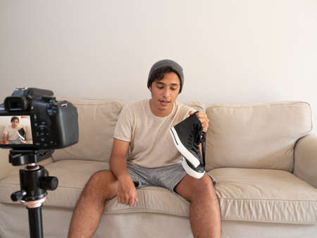 Hispanic young man creating clothing content for social networks with a camera on a tripod, sitting on a piece of furnitureの写真素材