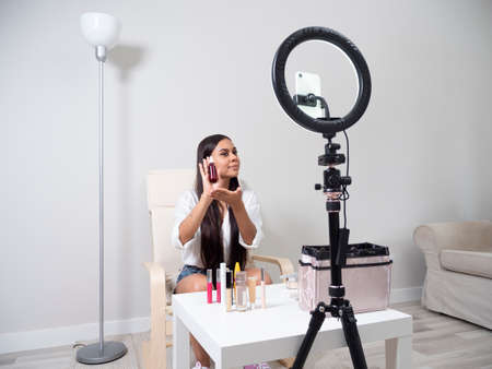 Young Latina content creator girl doing a makeup blog with different feminine products on a chair with a white table and a carpetの写真素材