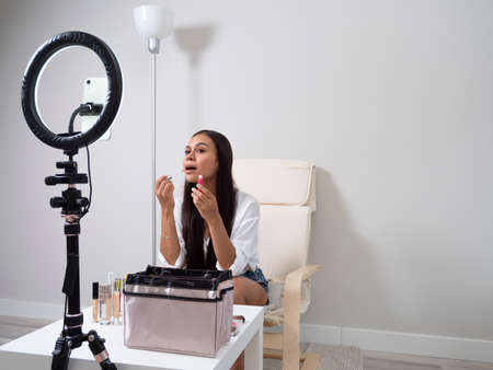 Young Latina content creator girl doing a makeup blog with different feminine products on a chair with a white table and a carpetの写真素材