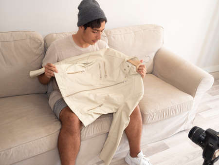 Hispanic young man creating clothing content for social networks with a camera on a tripod, sitting...