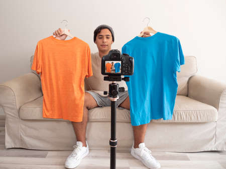 Hispanic young man creating clothing content for social networks with a camera on a tripod, sitting...