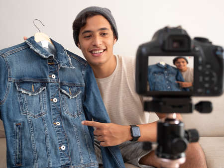 Hispanic young man creating clothing content for social networks with a camera on a tripod, sitting on a piece of furnitureの写真素材
