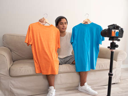 Hispanic young man creating clothing content for social networks with a camera on a tripod, sitting on a piece of furnitureの写真素材