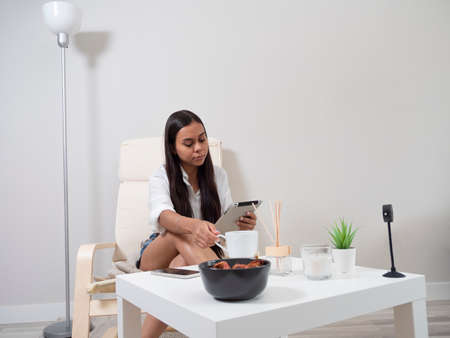 Young woman at home tired from work with a tablet sitting on a chair with a table in front of her in a relaxed atmosphere and drinking teaの写真素材