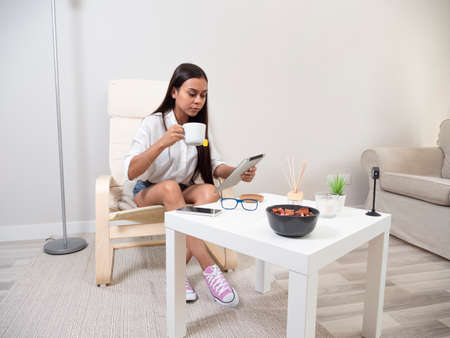 Young woman at home tired from work with a tablet sitting on a chair with a table in front of her in a relaxed atmosphere and drinking teaの写真素材