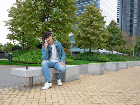 Latin man in a sweater and blue pants drinking coffee and talking on the phone in a place with many tall buildingsの写真素材