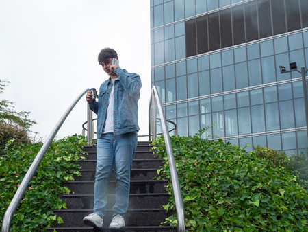 Latin man in a sweater and blue pants drinking coffee and talking on the phone in a place with many tall buildingsの写真素材