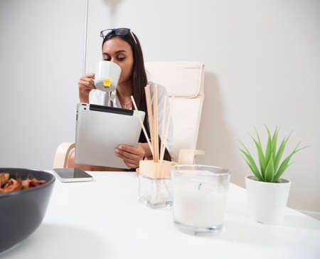 Young woman at home tired from work with a tablet sitting on a chair with a table in front of her in a relaxed atmosphere and drinking teaの写真素材
