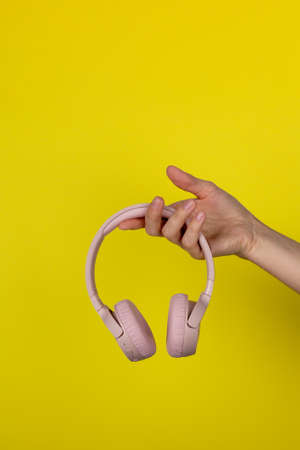 A young woman holds a pair of pink wireless headphones in her hand. Hand of a girl with headphones on a yellow background. Musical concept.の写真素材