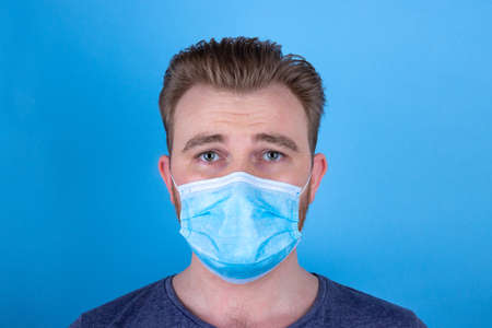 European man in a medical face mask on a blue background. Upset,confused,sad and disappointed man in a protective mask. A scared person wears a face mask to protect himselves and others from the virusの写真素材