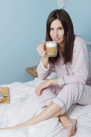 Smiling biracial young woman in pajamas sit on white bed drink hot coffee cappuccino in the morning....