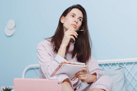 Portrait of brunette attractive cute creative cheerful girl student sitting in bed looking away making notes in notepad, using electronic gadget digital tablet in modern white light bedroom interiorの写真素材
