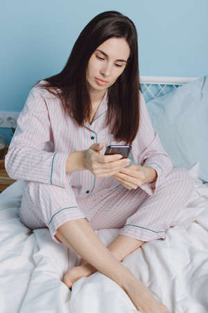 Close up view of brunette focused attractive millennial young woman using smartphone social media app sit on bed with white sheets. Relaxing at home holding phone texting message or play mobile gamesの写真素材