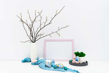 Blank photo frame for text, mockup with copy space for Easter holiday. Hand painted in trendy colors Easter eggs, geometric vase on tray with moss, feathers, branches, Easter bunny. Front view image.の写真素材