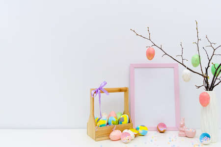 Trendy colorful Easter composition with hand painted eggs in yellow and grey colors in basket, geometric vase, confetti, twigs, pink Easter bunny. Blank photo frame, mockup with copy space for text.の写真素材