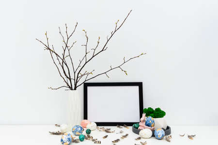 Blank photo frame for text, mockup with copy space for Easter holiday. Hand painted in trendy colors Easter eggs, geometric vase on tray with moss, feathers, branches, Easter bunny. Front view image.の写真素材