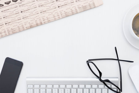 closeup of a business desk consisting on a financial newspaper, keyboard, mobile phone, wireless mouse, reading glasses and a cup of coffeeの写真素材