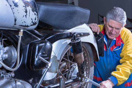 middle aged mechanic is tightening the shock absorber of a classic motorcycle with a wrench in process of restoration at his workshop - focus on the man faceの写真素材
