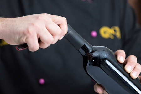 closeup of the hands of a young man uncorking a bottle of red wine cutting the closure with an...