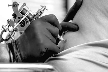 closeup of the hands of a tattoo artist tattooing the back of a young woman in the tattoo cabin at his tattoo shop - focus on the needleの写真素材