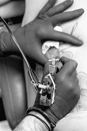 closeup of the hands of a tattoo artist tattooing the back of a young woman in the tattoo cabin at his tattoo shop - focus on tattoo machineの写真素材
