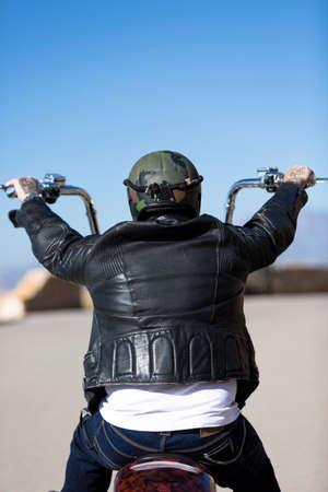 rear view of a chopper motorcyclist riding his customized motorcycle by a mountain road at sunrise -...