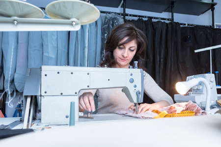 young seamstress is sewing the fabric seated on a sewing machine at her sewing atelier - focus on her right eyeの写真素材