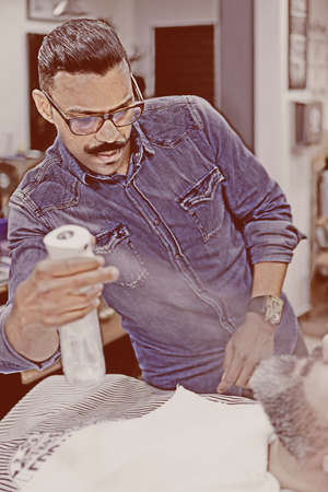 male barber is spraying the costumer's beard with a water spray on a beard shaving session in a barber shop - focus on the barber faceの写真素材