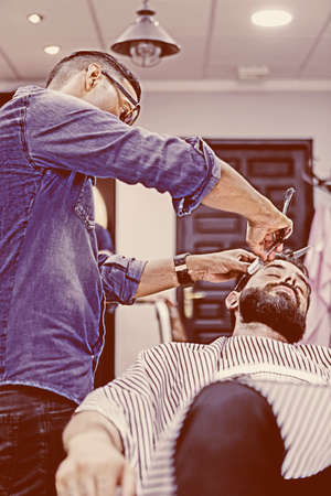 hairstylist on a beard shaving session and a customer on a barber chair on the barber shop - focus on the barber faceの写真素材