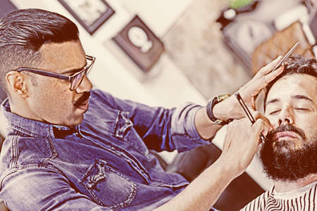 male hairstylist on a beard shaving session and a customer on a barber chair on the barber shop - focus on the barber faceの写真素材