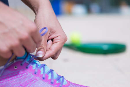 closeup of the hands of a woman is tying the shoelaces of the sneaker with a racket and a ball of...
