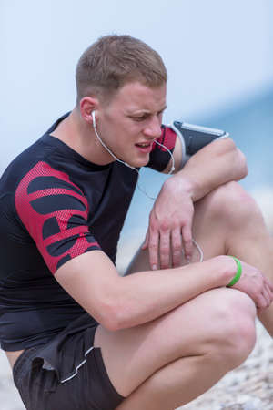 man runner is resting crouched while is listening music with headphones and a phone on an armband after a running session on the beach - focus on the frownの写真素材