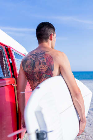 back view of a young man is holding a surf board next to a classic van on a surf session on the beach - focus on the girl tattoo faceの写真素材
