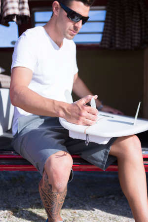 young man is preparing a surfboard sitting on a classic van on a surf session on the beach - focus on the thumbの写真素材