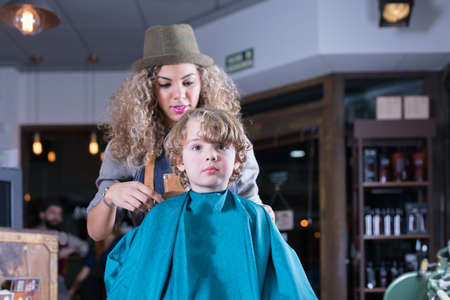Little boy sitting in barber chair, female hairdresser on background