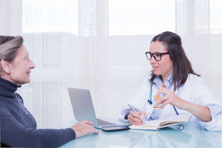 Young female doctor and old woman patient talking in consulting room while sitting at table.の写真素材