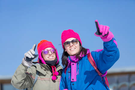 Smiling women in sunglasses and bright hats pointing at something on a ski resort