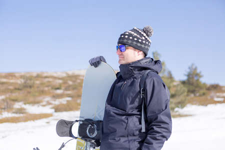 Smiling man in hat and sunglasses holding snowboard and looking away