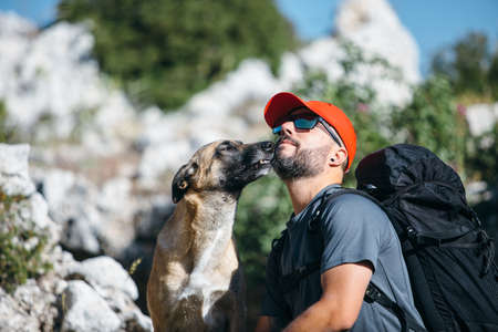 Portrait of pet kissing his bearded owner in red cap in sunlight at the mountainの写真素材