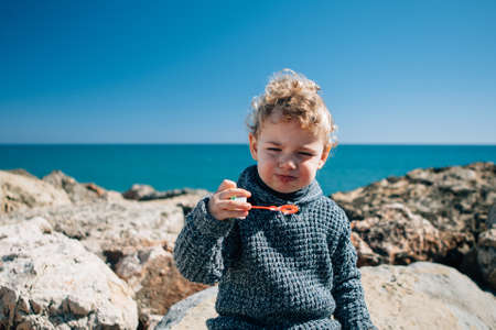 Portrait of blonde kid with windy hair blowing soap bubbles against of seascape
