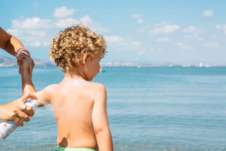 Back view of boy being sprayed by unrecognizable mother against of seascape. Copy space area availableの写真素材